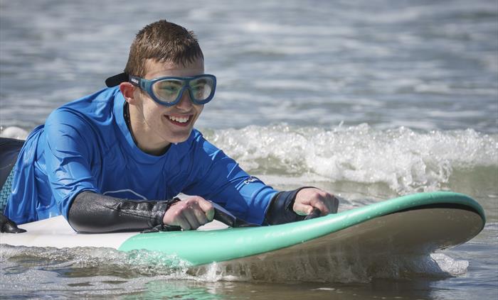 A close up of a young surfer who is disabled, smiling on a wave on a prone surfboard with goggles on
